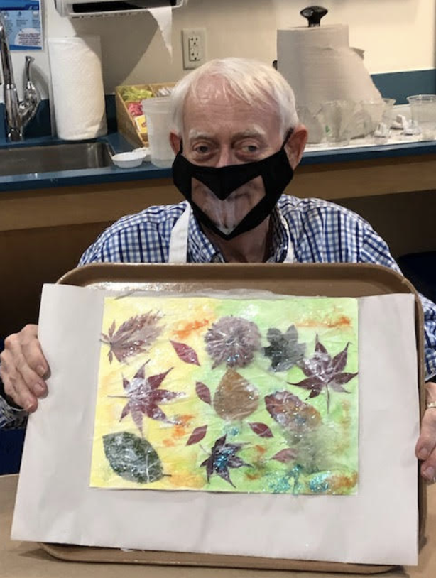 An older man holds up his art project featuring an array of leaves.