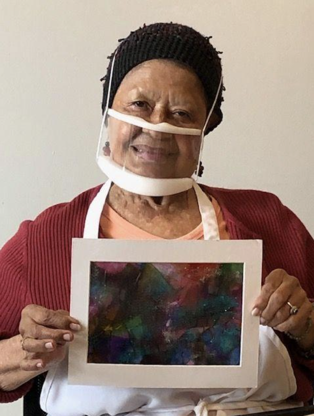 A woman holds up her finished art project, an abstract painting featuring dark tones.